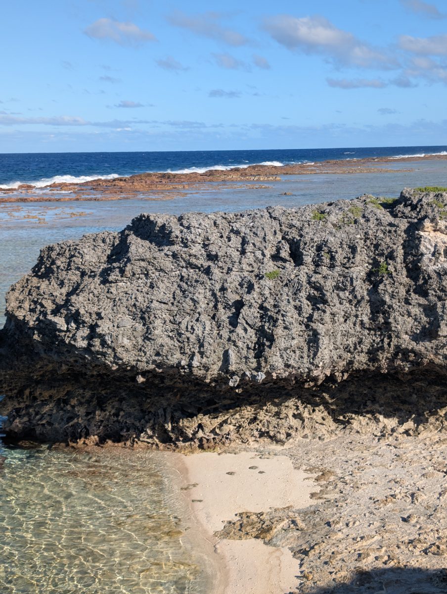Weathered undercut limestone spur on western Mauke above a shallow pool, with the exposed reef flat and breaking waves beyond.