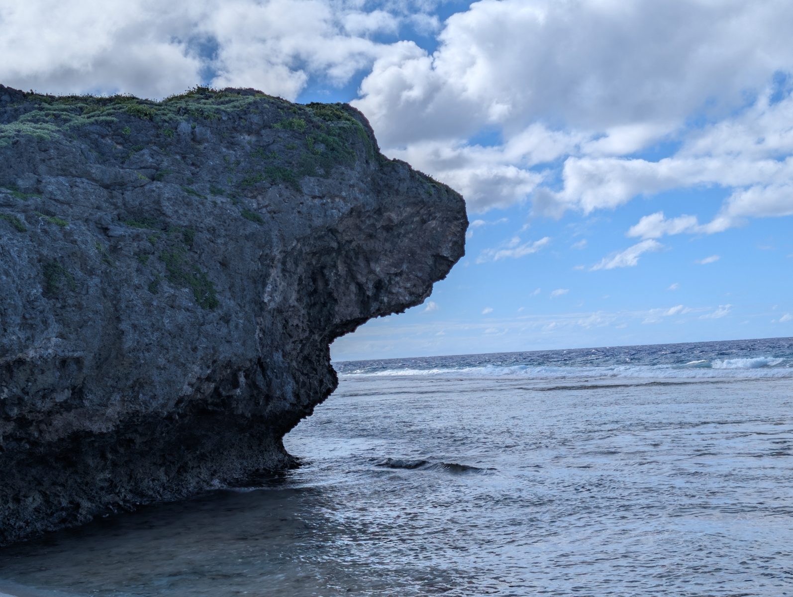 Deeply undercut limestone spur beside shallow reef-edge water, with the reef flat and breaking waves beyond at Arapaea.
