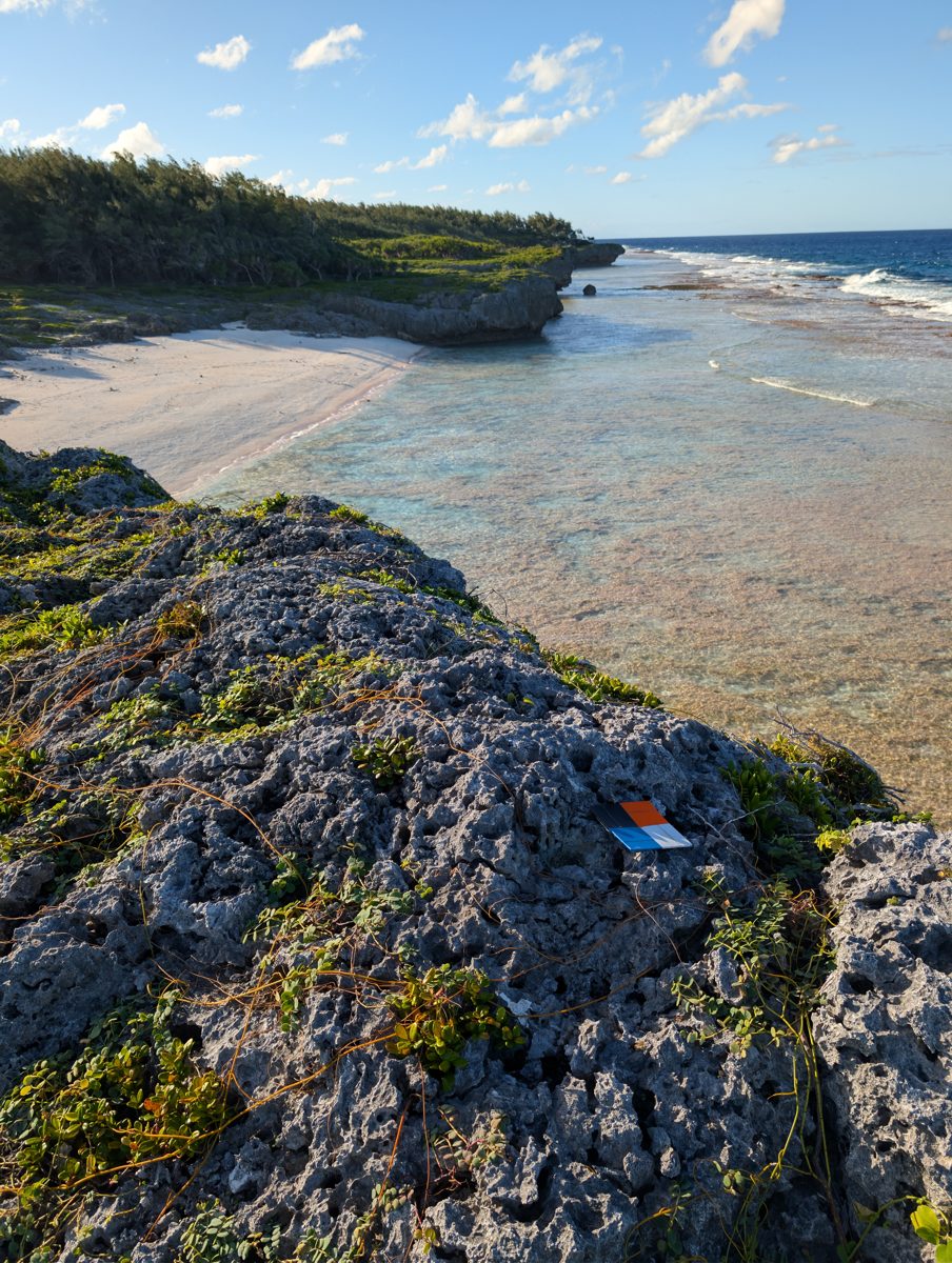 Marked survey target on rough coastal limestone above a narrow beach, shallow reef flat, and wave-cut coast at Aanga.