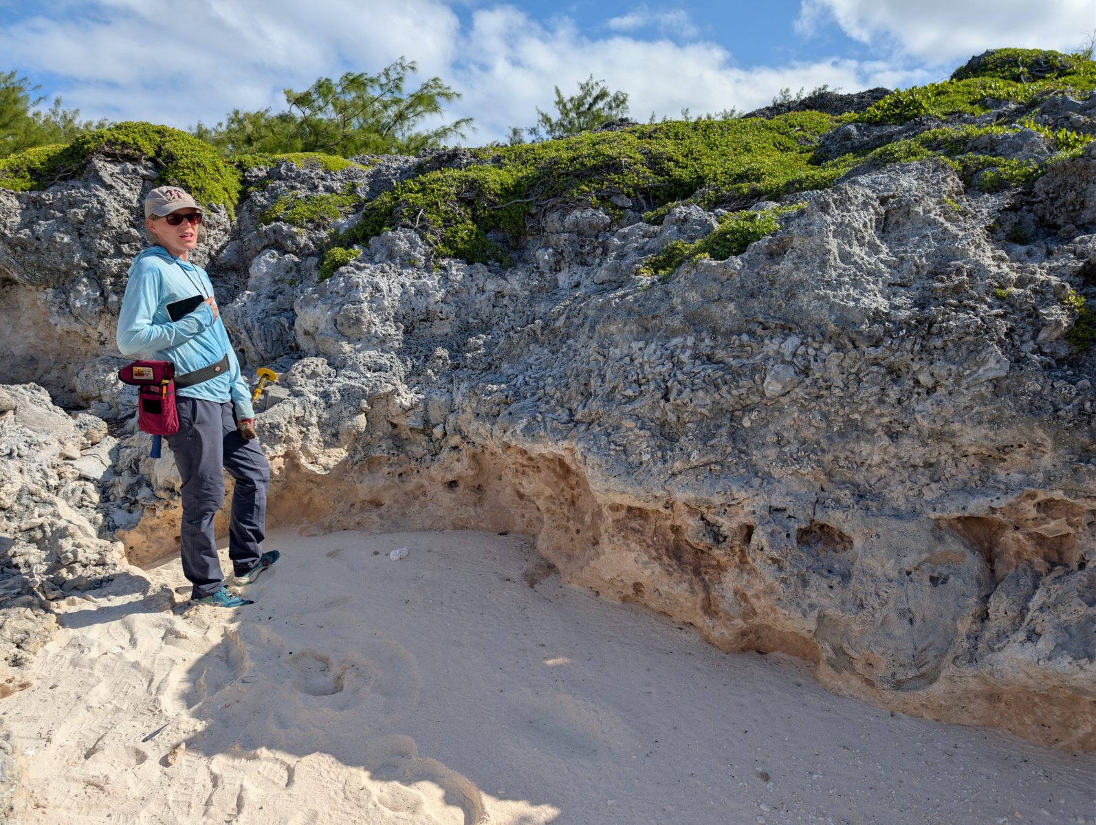 Jacky stands in an HS125 limestone groove beside a darker clast-rich band below rougher grey rock and low green vegetation.